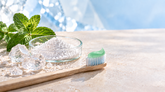 White mineral-like powder, mint and a toothbrush with toothpaste on a light surface.